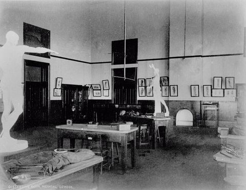 Black and white photograph. Two white statues of men in a classical style loom over long tables set with equipment in this empty, hall-like room.