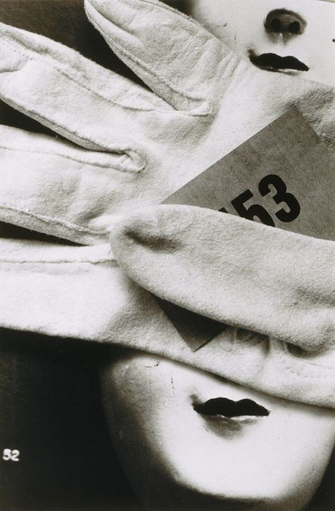 Black and white photograph. An empty white glove has a piece of paper printed with the number “53” tucked under the thumb. The glove fills the top two-thirds of the picture. Two pale doll’s faces with dark, smiling lips are seen from the nose down. There is one in the upper right corner and one in the corner below.