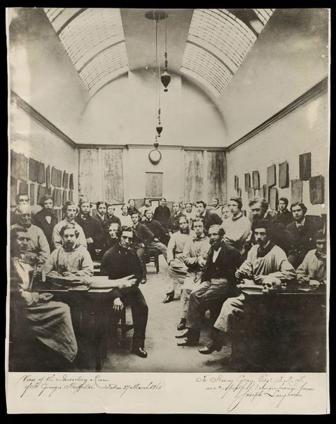 Black and white photograph. About forty men pose in a hall-like dissecting room. The ceiling is rounded, like a tunnel, and set with skylights. A pair of feet hang off the end of the table closest to us to our left and a skull sits at the front edge of the table to our right.