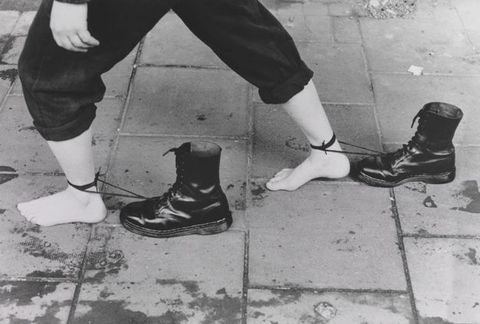 Film still. Shown from the hips down, a barefooted person walks on a sidewalk in this black and white image. One foot is planted far in front of the other, and each is tied around the ankle with the long laces of distinctive high, black Doc Martens boots.