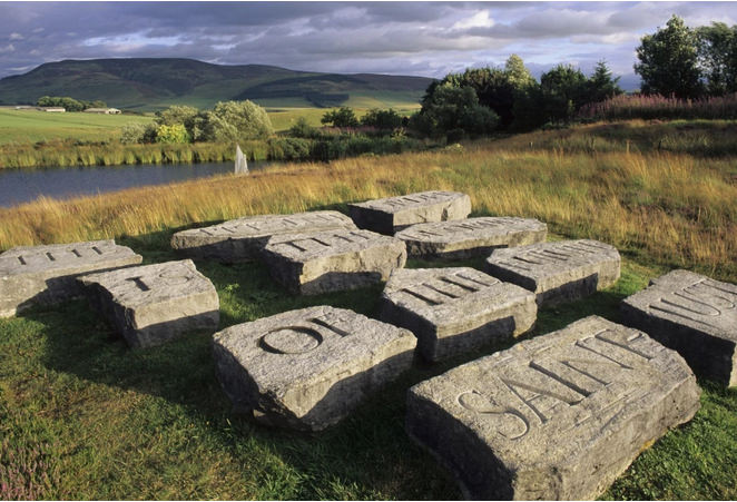 Sculpture. Eleven rectangular or square grey stones are each carved with a single word in all caps. The stones lie in a grassy field near a river.