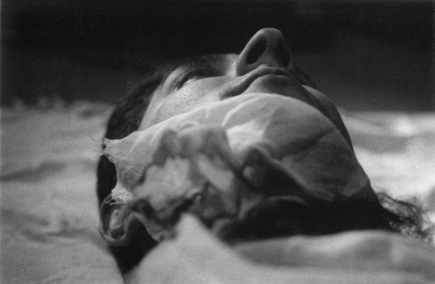 Black and white photograph. The underside of a man’s chin and nose is taken as if the camera was placed on his shoulder or chest. A piece of fabric seems caught up under his chin. His nose is like a peak rising over the mountain range of his face.