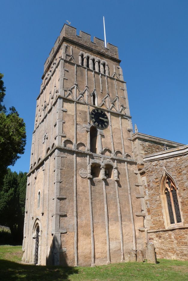 Architecture. From across a grassy lawn, we look up at a square tower. Four levels get narrower the higher up they go to make up the façades. A black clock with gold numbers is about halfway up the tower on the side closest to us. A stone structure is built to adjoin the tower to our right.