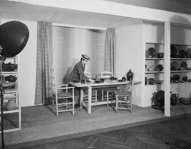 Black and white photograph. On a low platform in an exhibition hall, a pale-skinned woman arranges dark bowls and mugs on a dining table. A false window is hung with curtains behind the table, and shelves are lined with pots and plates to either side. An industrial-style spotlight illuminates the scene from our left.
