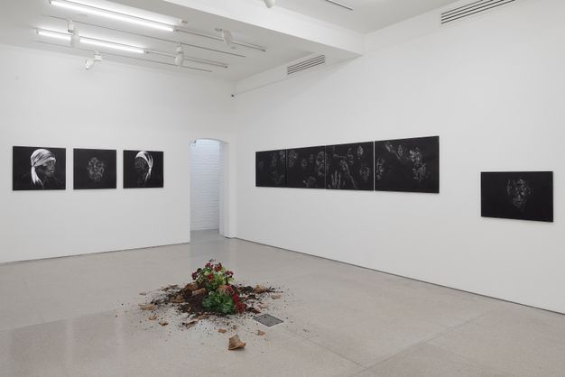 Installation photograph. Three terracotta pots, along with the dirt and red-flowered plants they held, lie broken in a heap at the centre of a gallery hung with black and white chalk drawings of faces. The drawings are done in white against black paper so have an ethereal look.