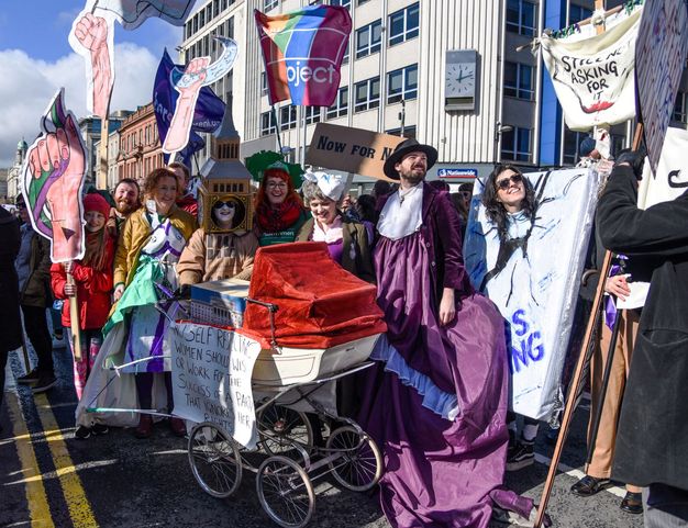 Colour photograph. Smiling men and women, all with pale skin and most in costumes, hold up or look towards signs. For instance, one person is dressed up as Big Ben, her white face smiling through the open clock face. She stands behind a pram hung with a handwritten sign that read, “No Self Respecting Women Should [indecipherable] or Work for the Success of a Party that Ignores Her Rights”.